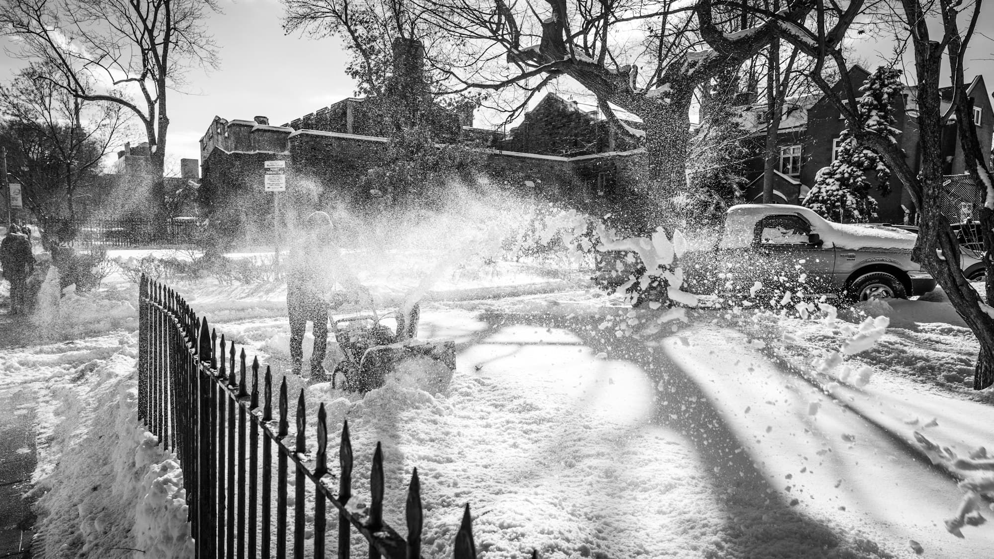 Snowblower clearing a sidewalk beside parked cars on a residential street